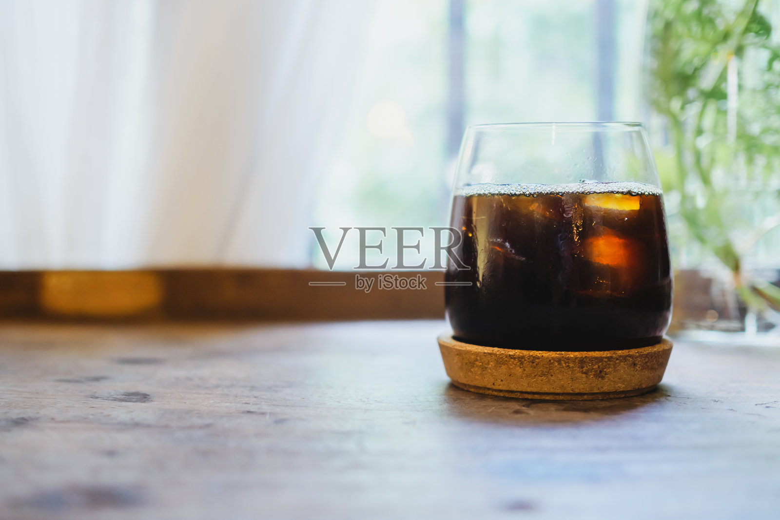 木桌上的玻璃杯里盛着冰美式咖啡,白色窗帘和窗底照片摄影图片