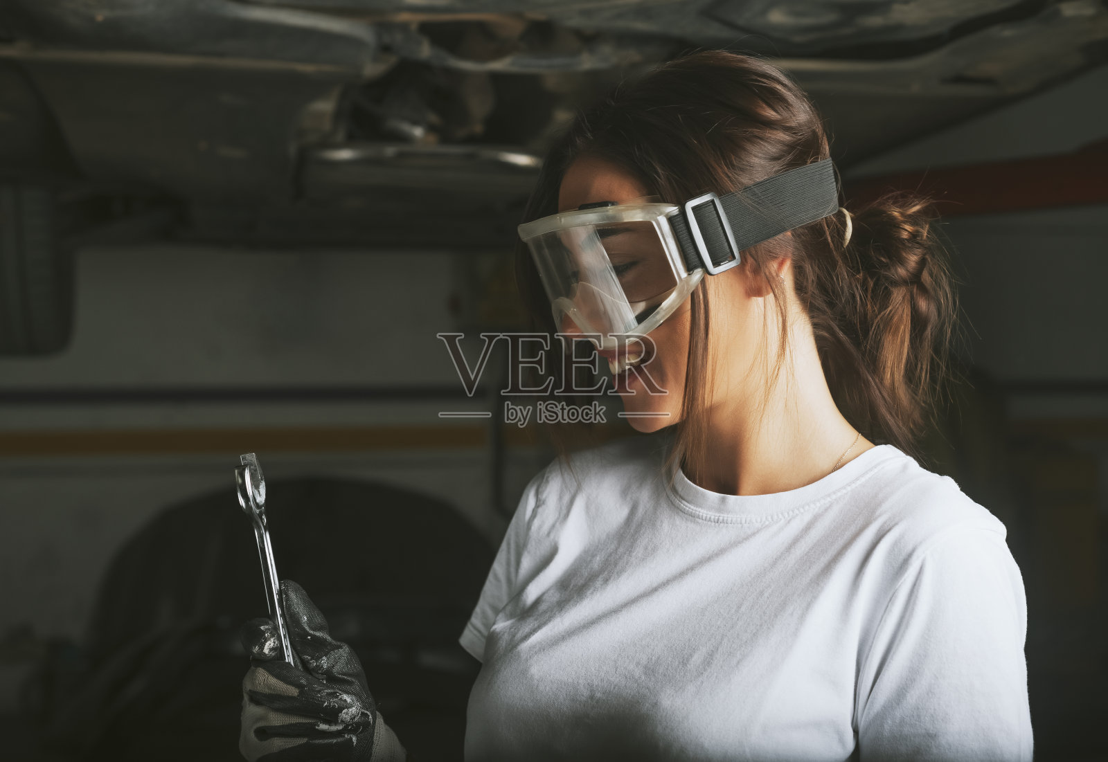 一位带着护目镜微笑的年轻女机械师正在寻找修理汽车的工具。照片摄影图片