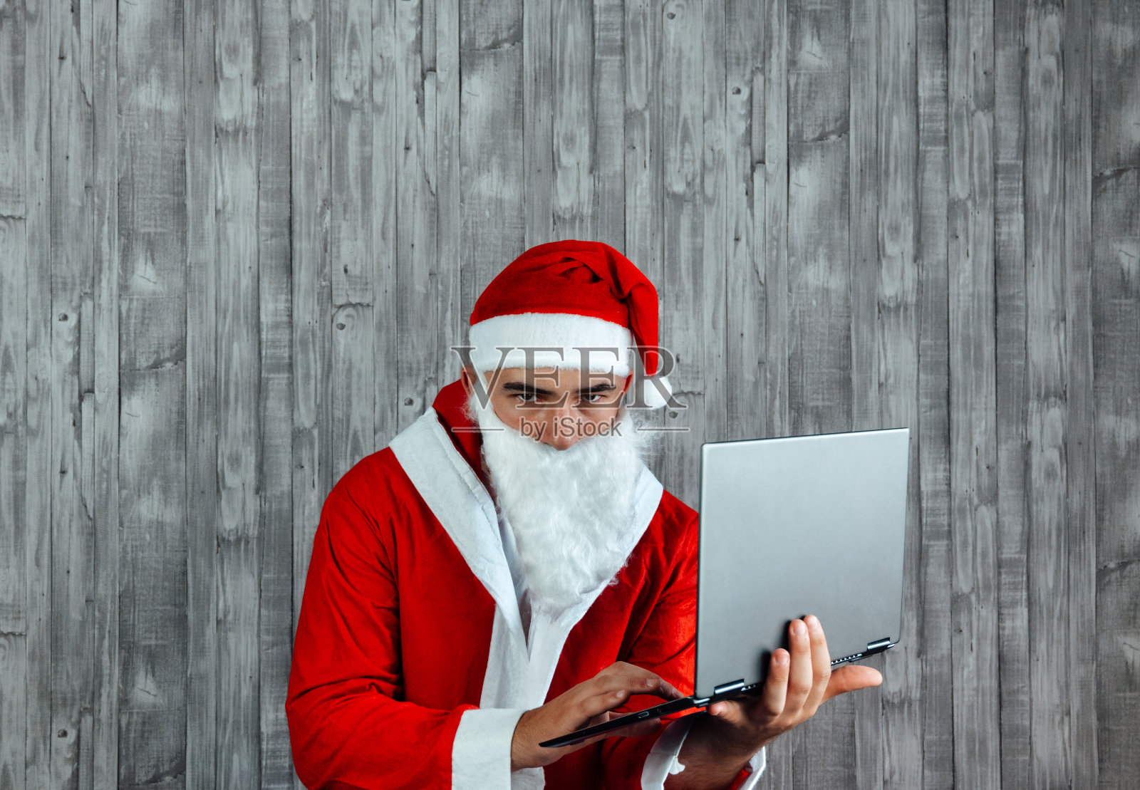 白人年轻男子打扮成圣诞老人,手里拿着笔记本电脑生气。圣诞节的销售理念照片摄影图片