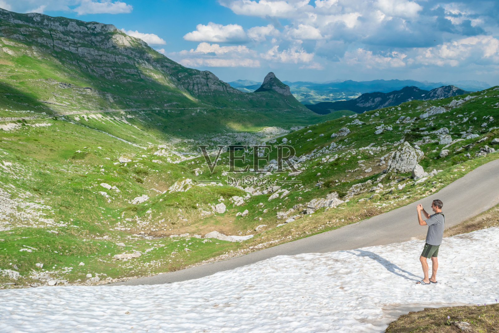 一名游客站在雪地里拍摄美丽的夏日风景。Durmitor国家公园。黑山最高的公路隘口。照片摄影图片