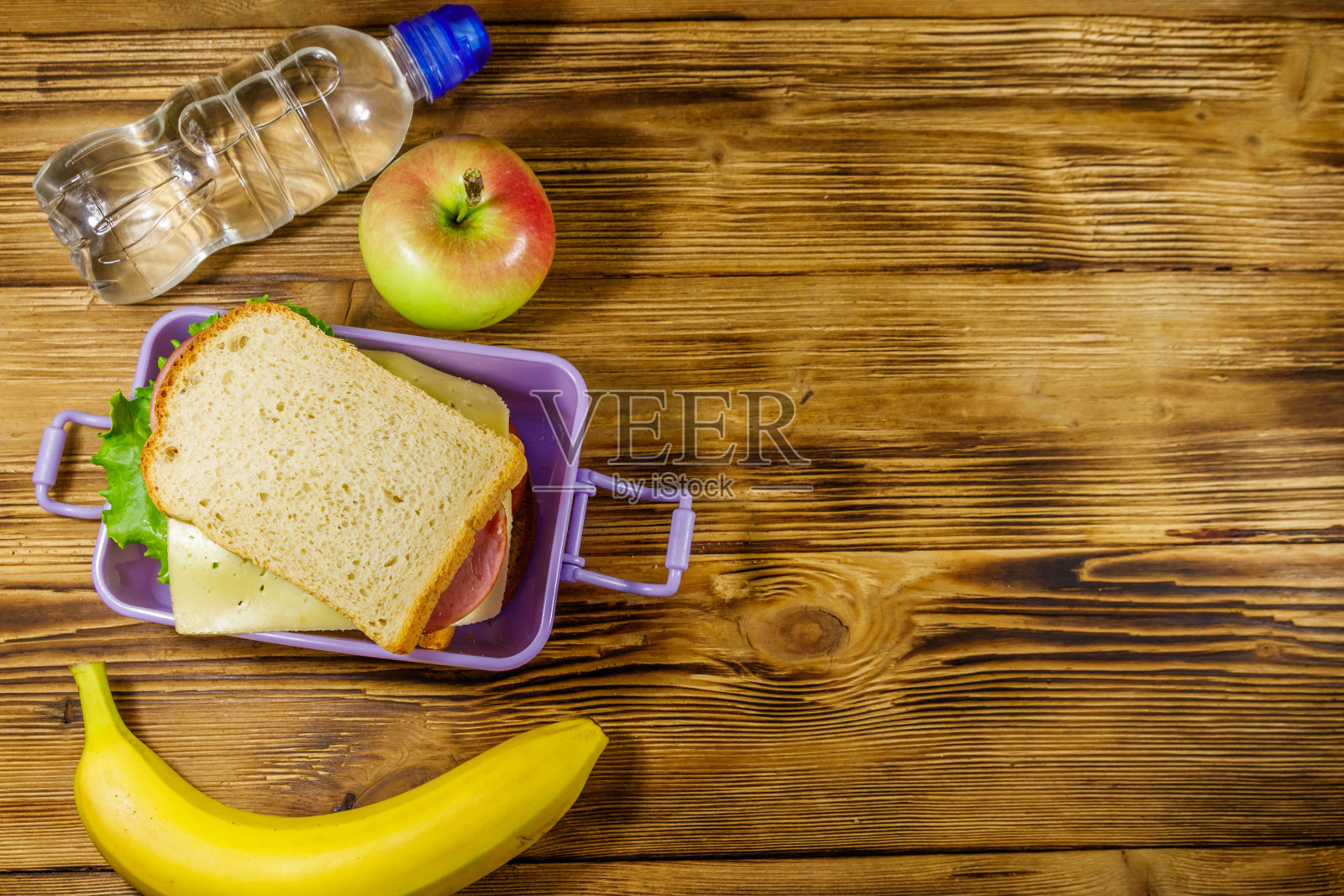 木桌上放着三明治、一瓶水、香蕉和苹果的午餐盒。俯视图,复制空间照片摄影图片