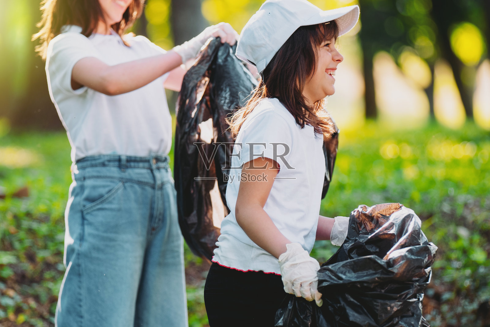 侧视图照片,一个快乐的小女孩戴着白色的帽子和t恤,戴着手套,把垃圾袋放在垃圾里,其他女孩站在旁边,拿着垃圾袋,保持大自然的清洁。照片摄影图片