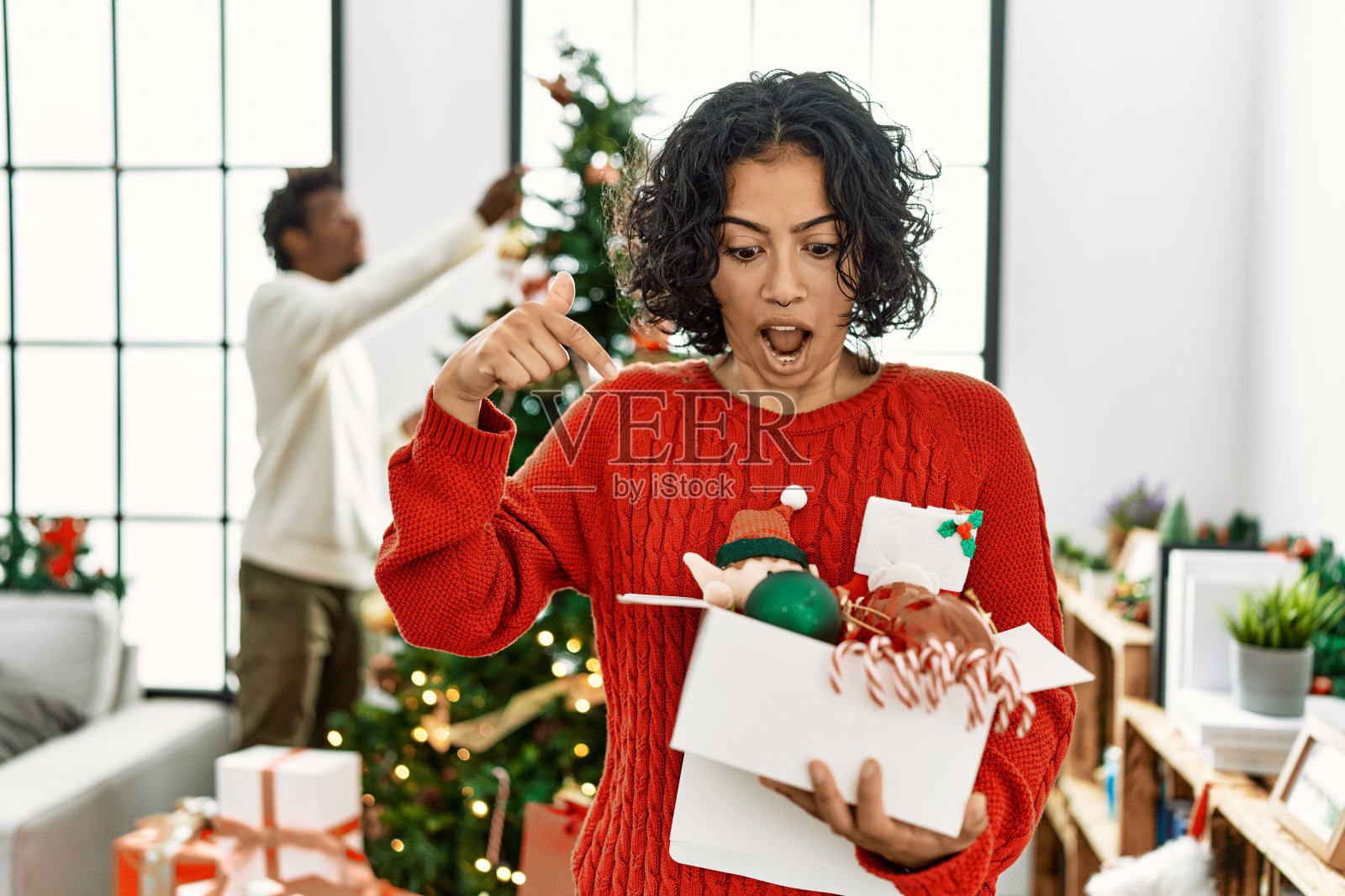 年轻的西班牙妇女站在圣诞树旁,她的手指向下指着广告,惊讶的脸和张开的嘴照片摄影图片