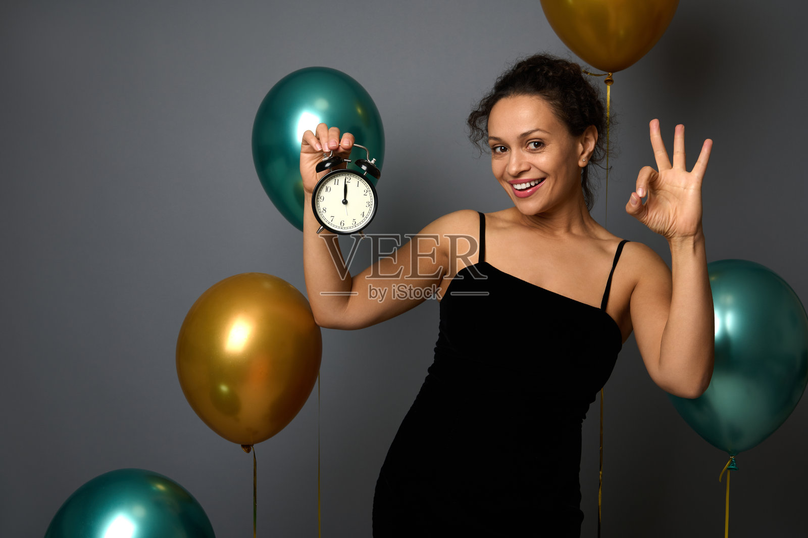 快乐的混血儿女子手持闹钟,午夜,显示OK标志微笑看着相机,孤立在灰色背景气球。新年快乐,圣诞快乐广告理念照片摄影图片