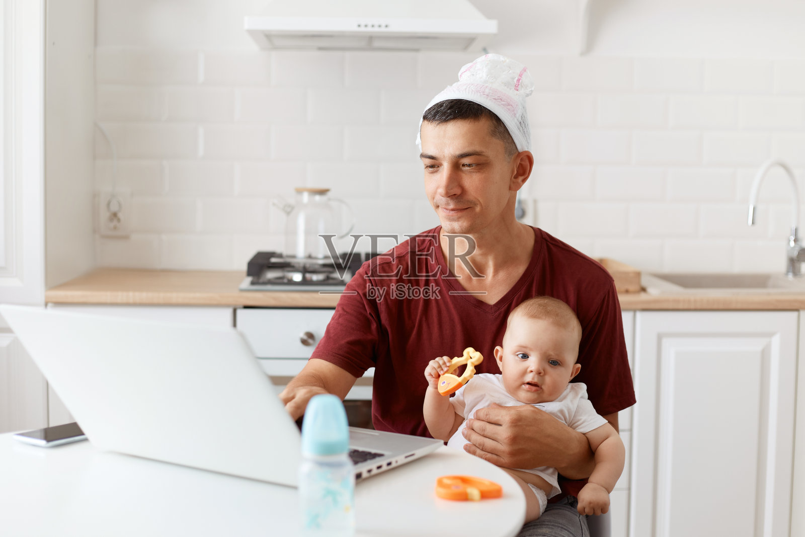 有趣的集中英俊的自由职业者男性肖像穿着紫红色t恤,在白色厨房摆姿势,坐在笔记本电脑前,手里拿着婴儿和工作。照片摄影图片