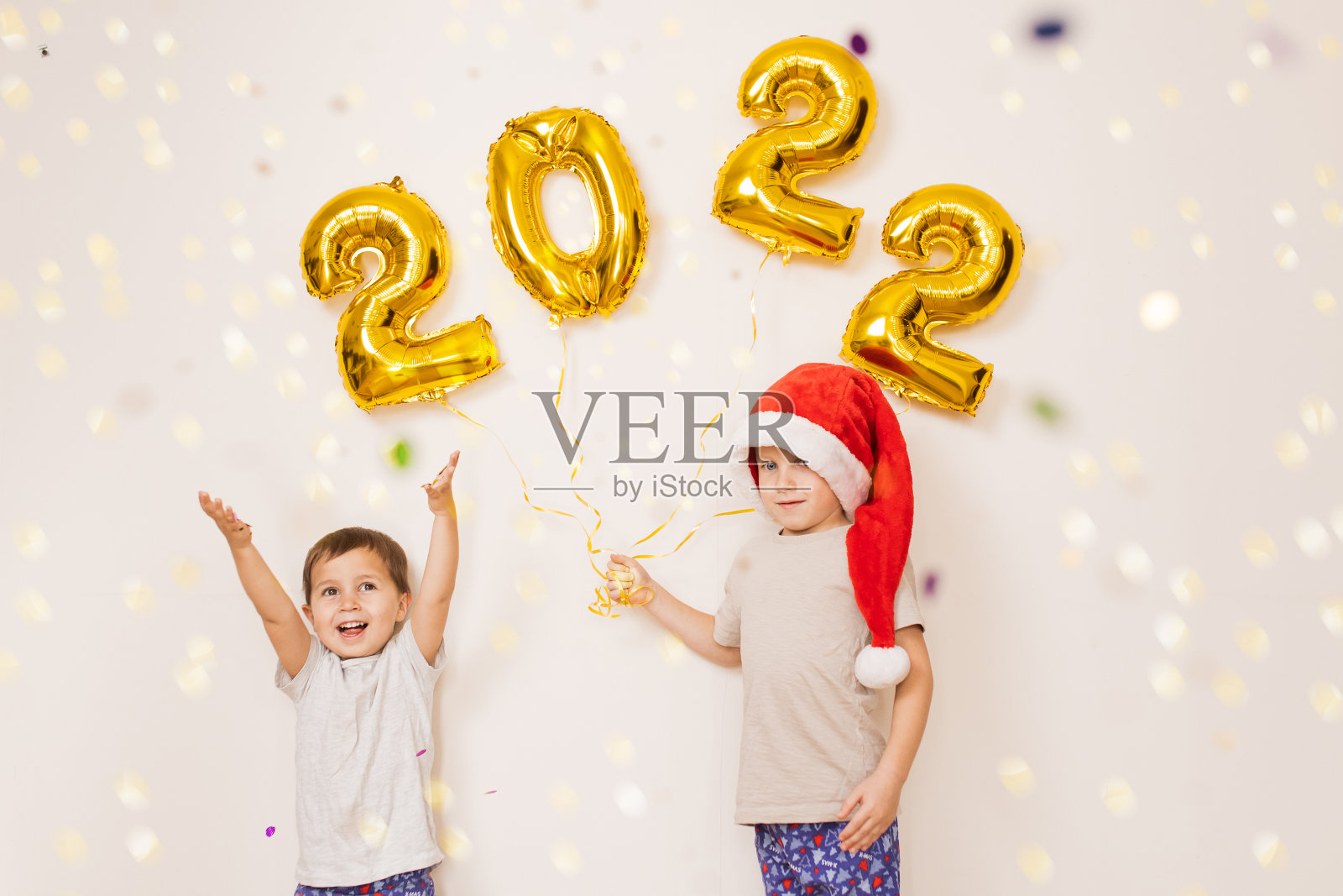 孩子们戴着红色的圣诞帽,开心地微笑着,在挂着金色气球的白色墙壁旁,享受着五彩纸屑。在家里庆祝2022年新年。圣诞节的概念照片摄影图片
