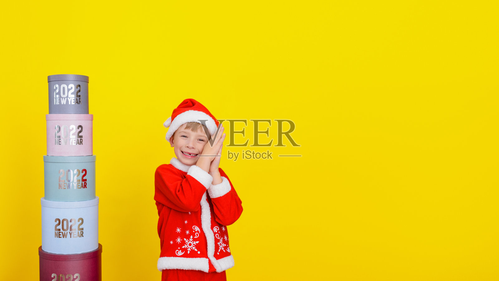 一个六岁的可爱的孩子站在一个圆圆的礼品盒旁边,上面写着“新年2022”,他笑得很开心,黄色的背景,复制空间。照片摄影图片