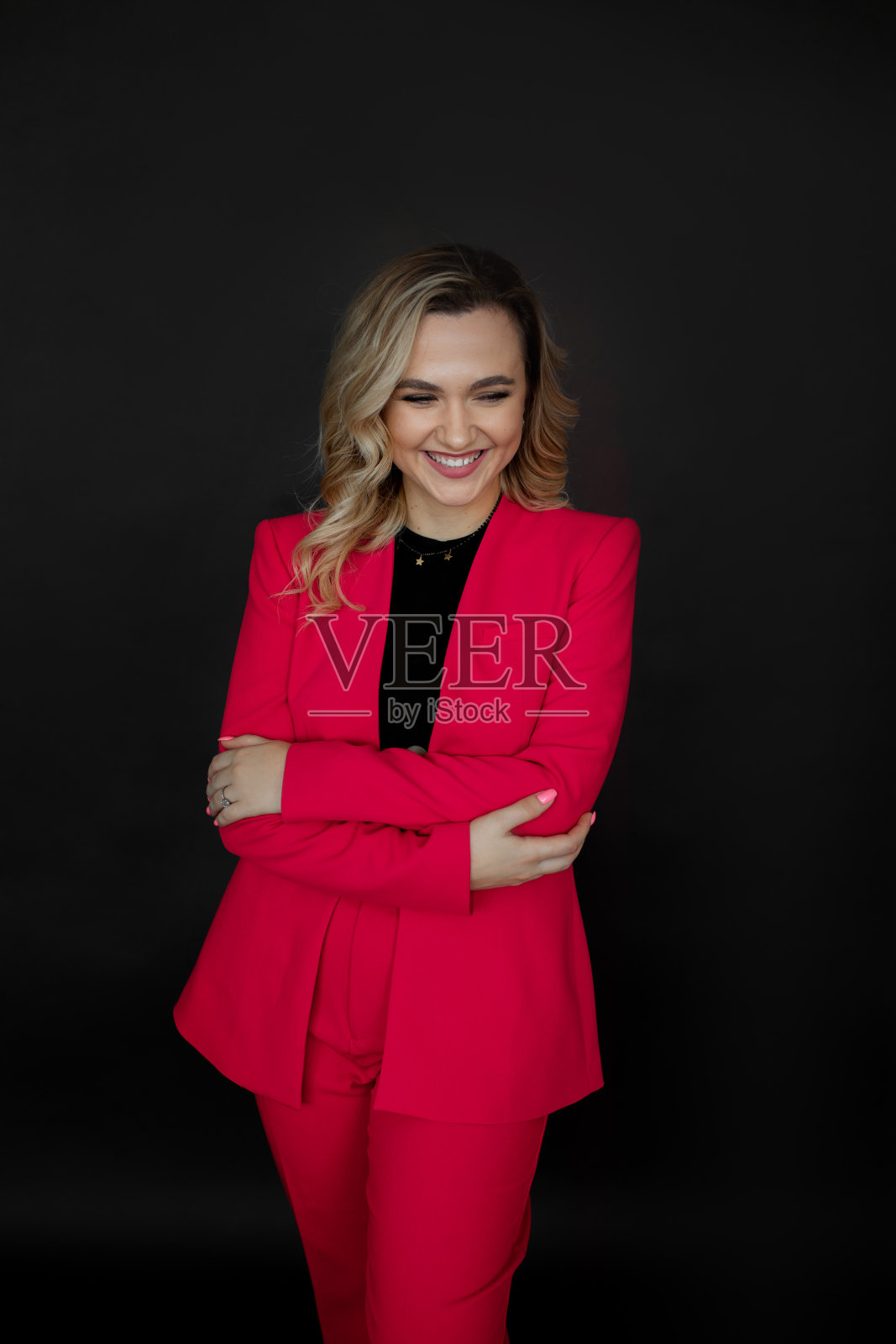 一个可爱的年轻金发女人,微笑着看着镜头,在摄影棚里,穿着亮红色的长裤套装,在黑色背景下摆姿势。照片摄影图片