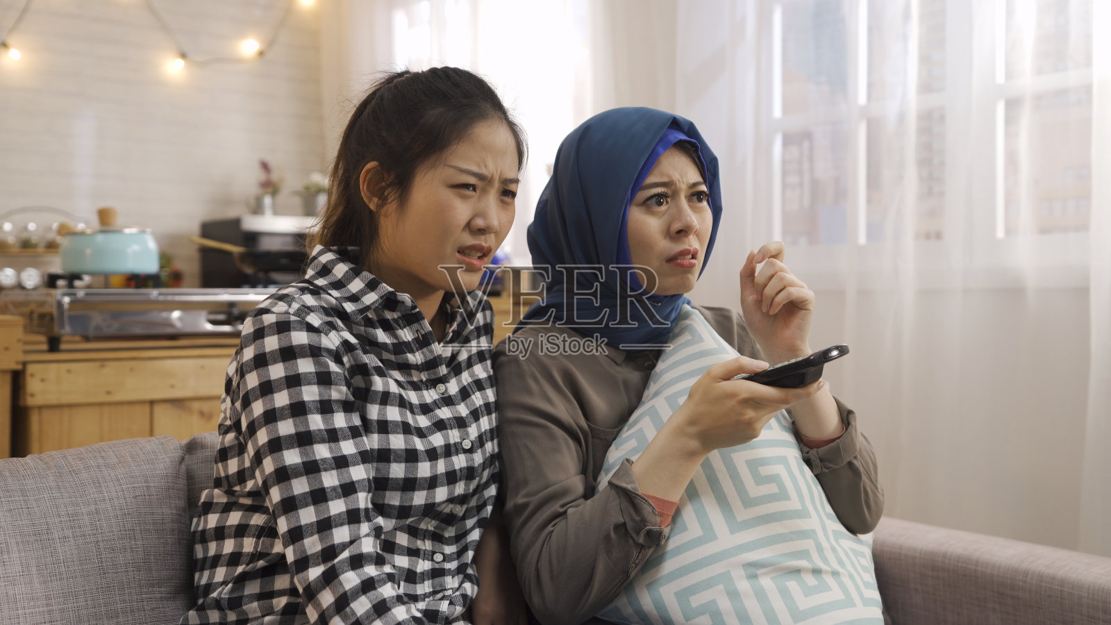 各种各样的女性最好的朋友坐在沙发上皱着眉头,拿着电视控制器看电视上的恐怖电影。两个亚洲阿拉伯和日本室友被惊悚电影的拥抱枕头吓到了照片摄影图片