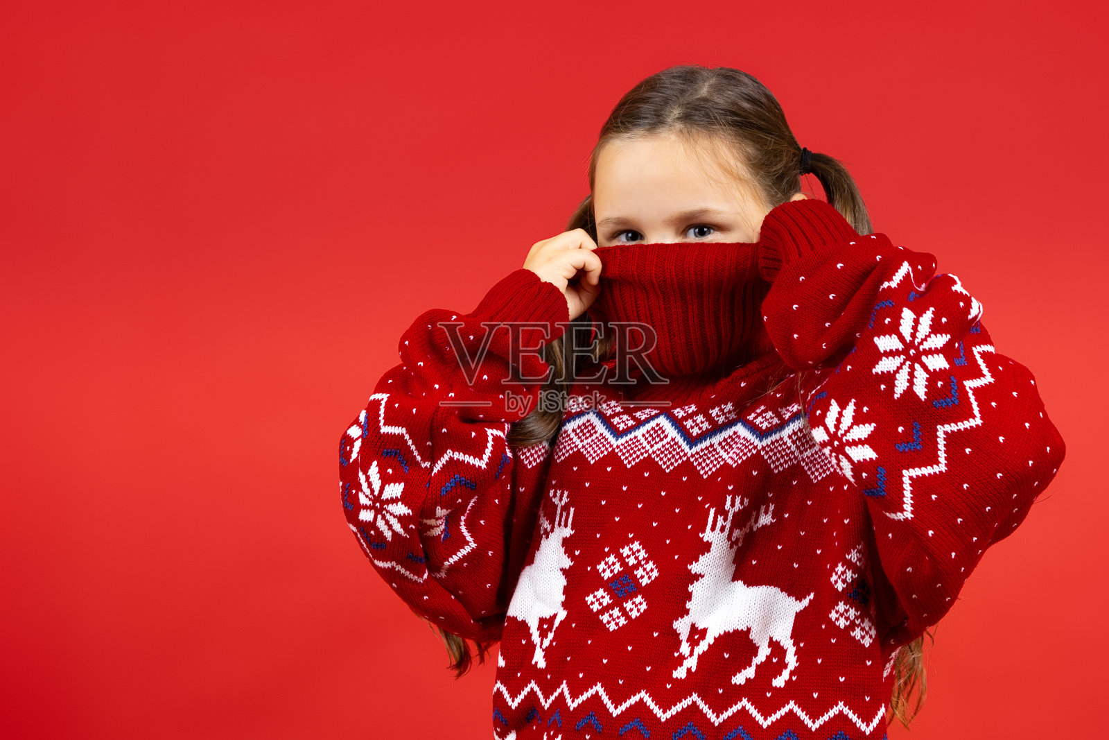 顽皮女孩的近距离肖像藏在红色圣诞毛衣与驯鹿的半脸,孤立在红色背景照片摄影图片