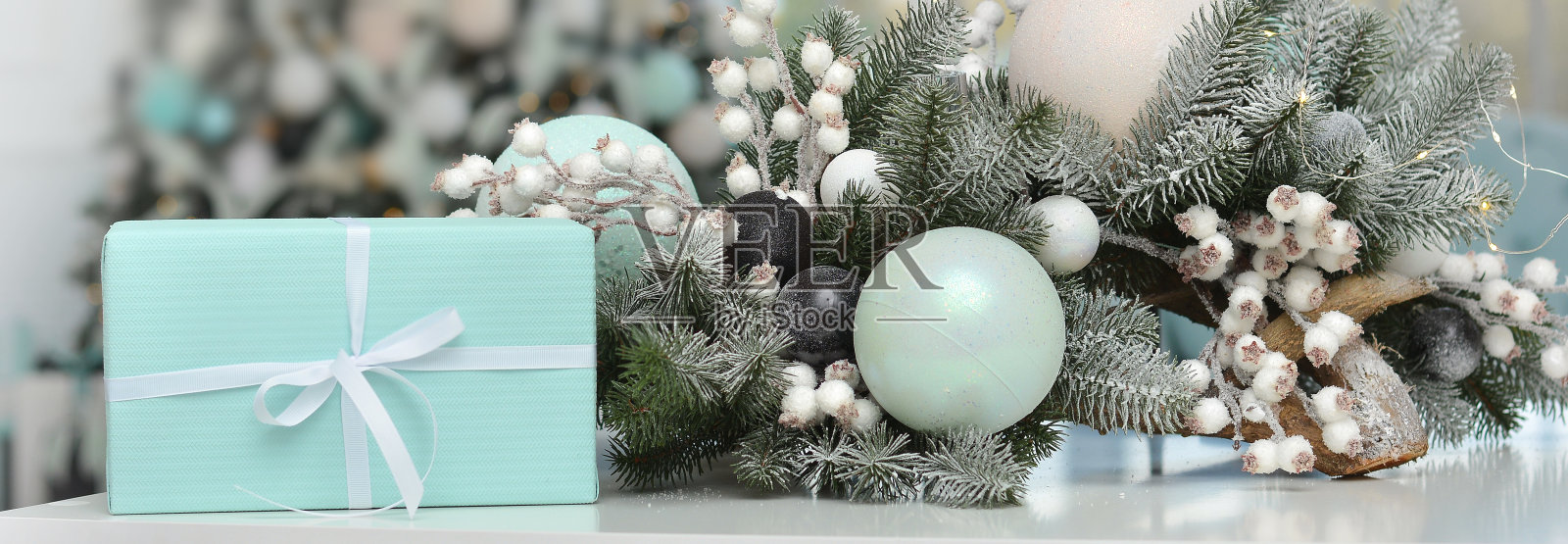 蓝色的盒子旁边放着一份美丽的圣诞礼物,上面有白雪覆盖的圣诞树树枝,新年的球,白雪覆盖的浆果。横幅照片摄影图片