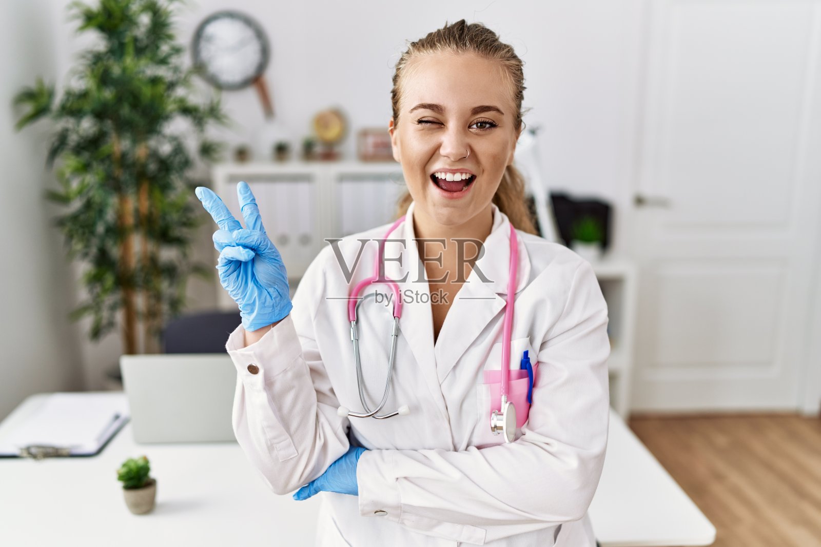 年轻的白人女性穿着医生制服,戴着听诊器在诊所里微笑着,对着摄像机眨着眼睛,做着胜利的手势。二号人物。照片摄影图片
