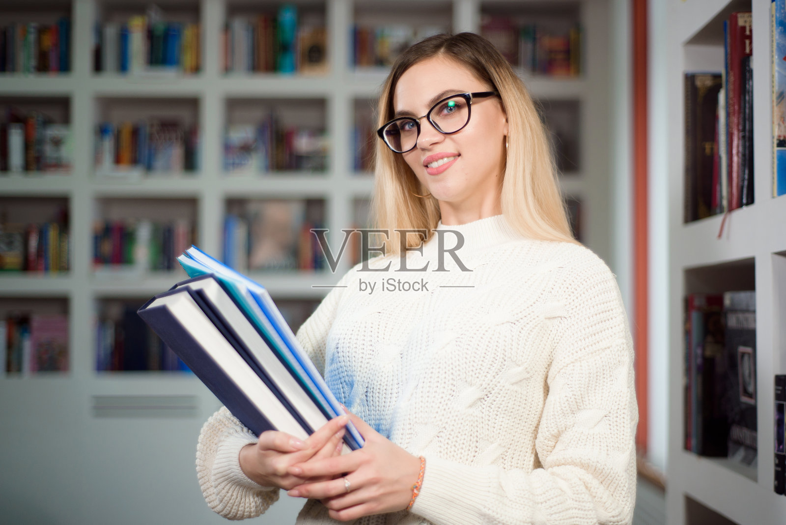 开朗的女学生,戴着眼镜,站在现代室内图书馆的书架旁。照片摄影图片