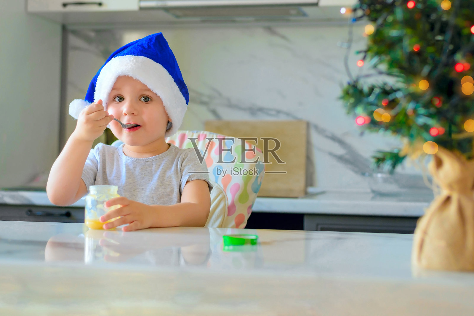 戴着圣诞帽的搞笑男孩在厨房里吃罐子里的苹果和梨泥。婴儿食品的概念。照片摄影图片
