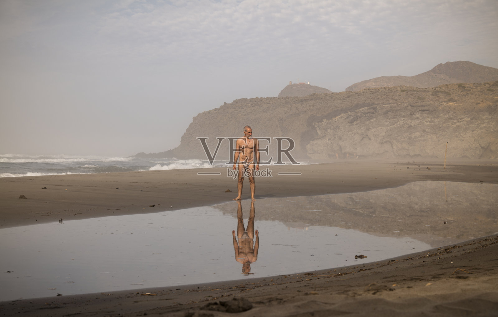 上身赤裸的男子泳装站在风景优美的海滩上,倒影在水面上,在卡波德加塔自然公园,阿尔梅里亚,西班牙照片摄影图片