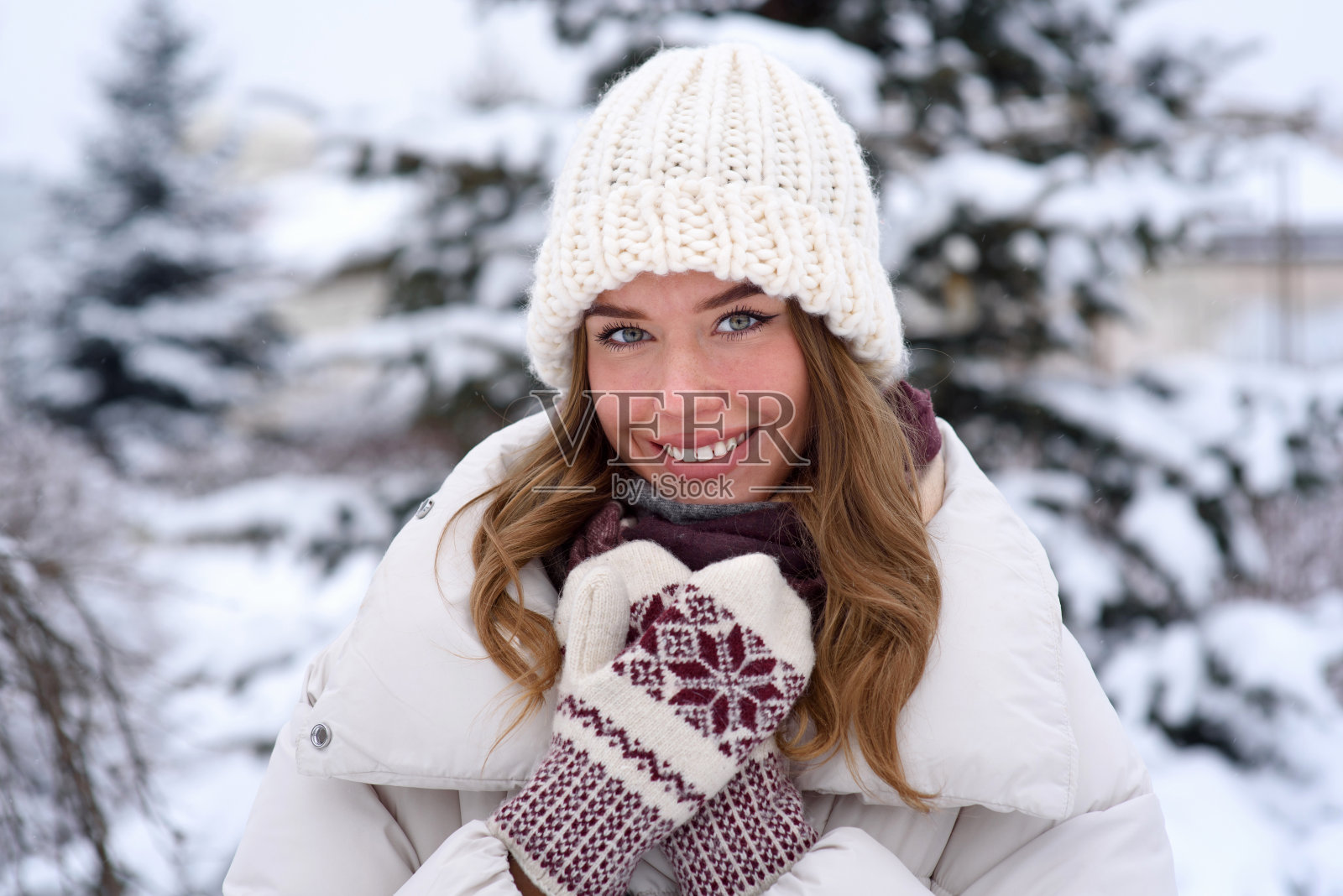 一位面带微笑的年轻女子的肖像,戴着针织帽,手套和暖和的夹克,在冬天积雪覆盖的公园里散步。照片摄影图片