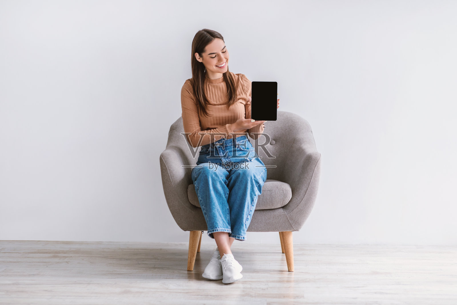 快活的年轻女士坐在扶手椅,显示平板电脑与空屏幕对白墙,为网站或应用程序的模型照片摄影图片