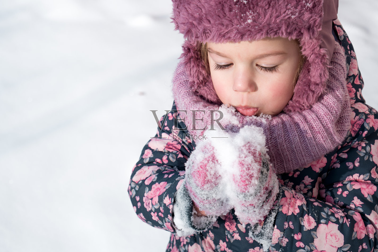 冬天,游戏,家庭,童年的概念-特写肖像真实的小学龄前小女孩在粉红色的帽子温暖的衣服有乐趣的微笑在寒冷的霜冻天气。有趣的孩子在白雪上吹气照片摄影图片