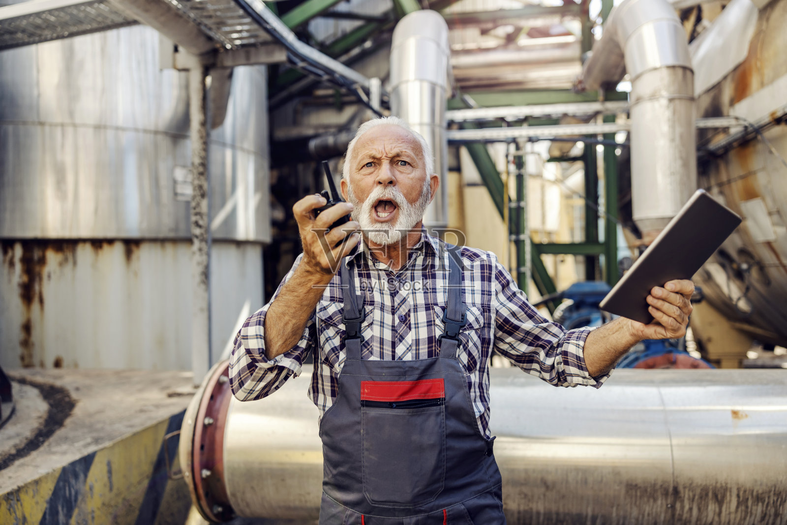 一位愤怒的高级主管站在工厂里,手里拿着一个平板电脑,对着对讲机大喊。没有时间和地点犯错。愤怒的主管对他的员工大喊大叫。照片摄影图片