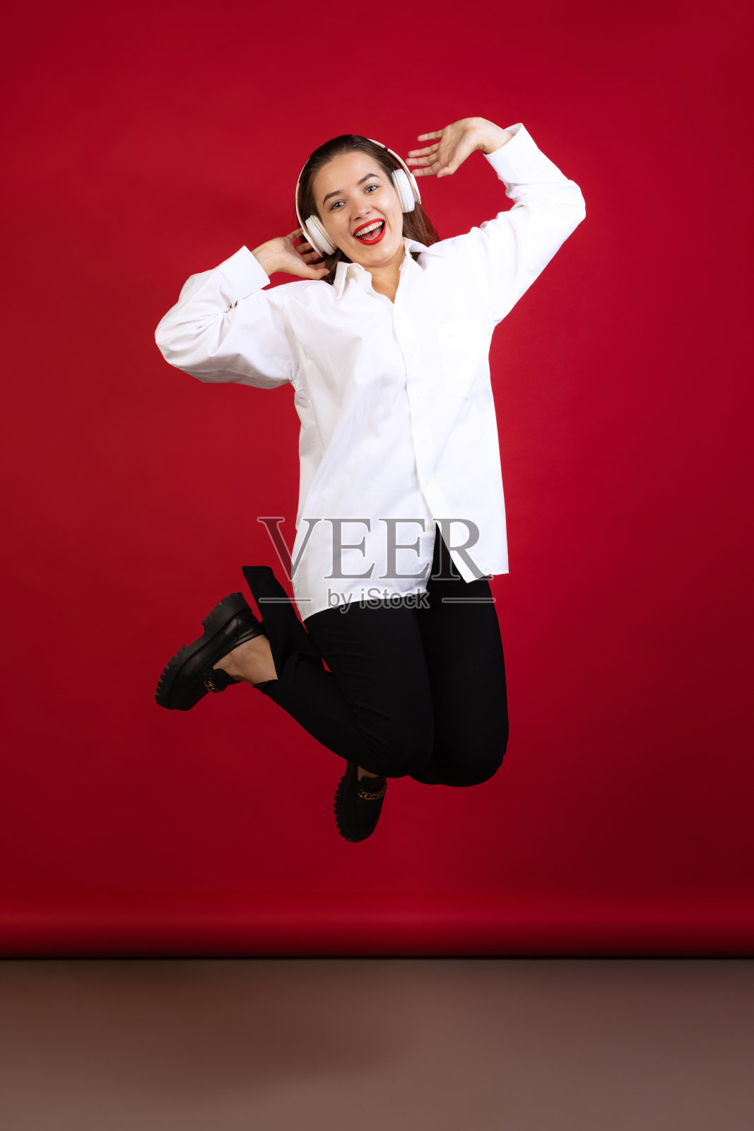 乐观的年轻美丽的女孩穿着白衬衫笑和跳跃孤立在暗红色的演播室背景。情感的概念,时尚照片摄影图片