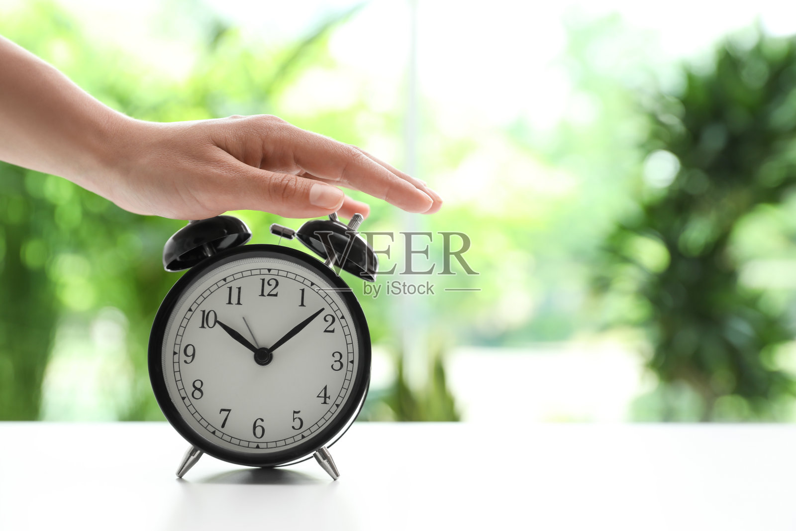 在模糊的背景下,白色桌子上的女人关掉黑色闹钟,特写。空间的文本照片摄影图片