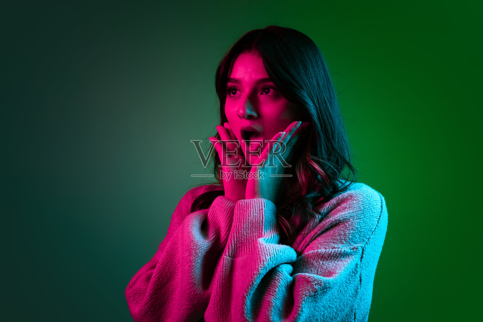 工作室拍摄的年轻可爱的女孩,学生穿着休闲风格的衣服在暗绿色的背景下,在粉红色的霓虹灯下孤立地摆姿势。照片摄影图片