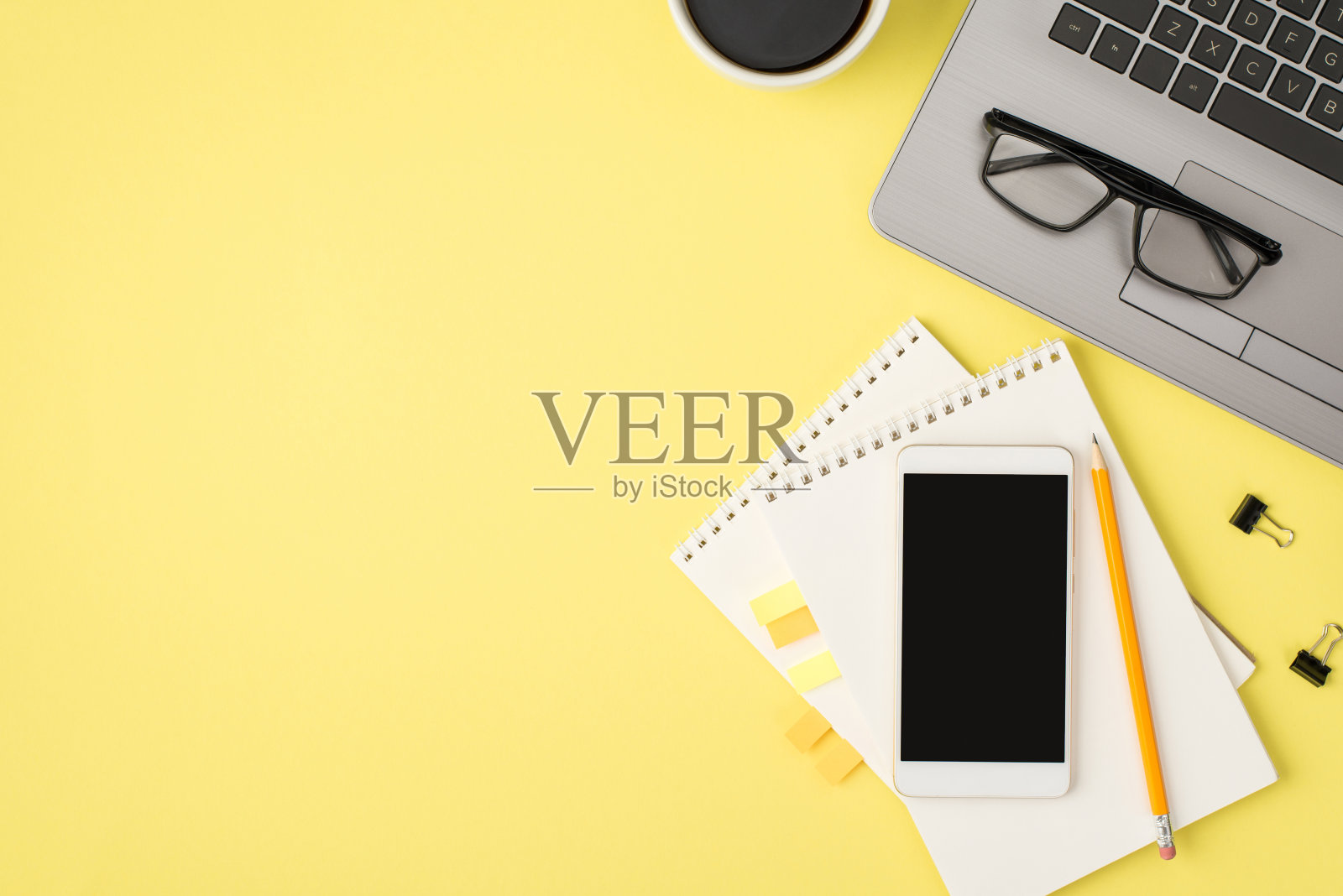 俯视图照片的眼镜在笔记本电脑,杯子,饮料,铅笔装订夹,智能手机和两个螺旋记事本与黄色塑料粘合剂书签在孤立的淡黄色背景与copyspace照片摄影图片