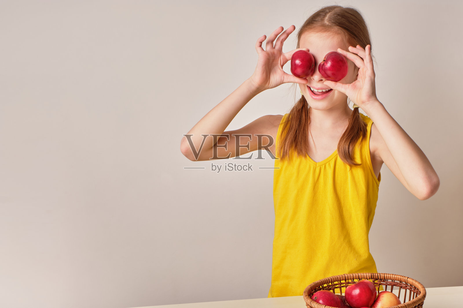 一个积极好玩的女孩与苹果在灰白色的背景。一个快乐的孩子,一个微笑的女孩,手里拿着新鲜的成熟的红苹果。附近有一篮子苹果。健康有机营养的概念照片摄影图片