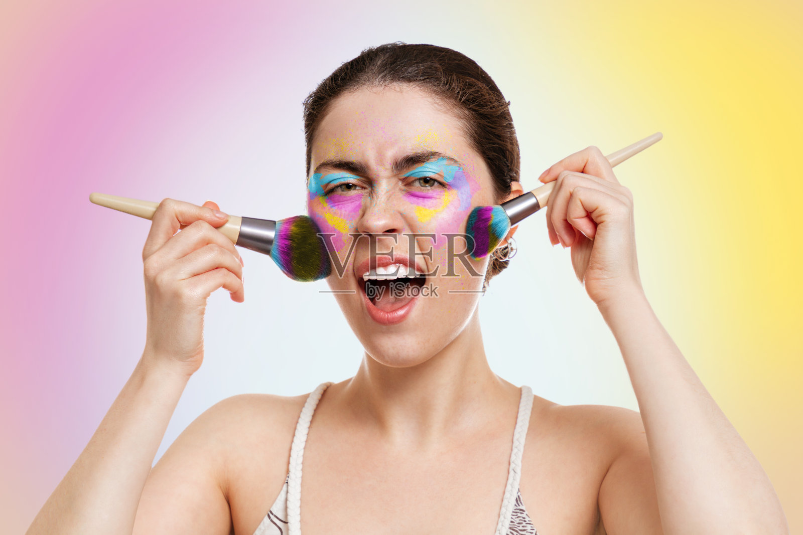 一个年轻漂亮的女人,画着明亮的眼睑妆,拿着化妆刷靠近脸颊,尖叫着。Yellow-pink背景。副本的空间。化妆、自我护理和化妆品的概念照片摄影图片
