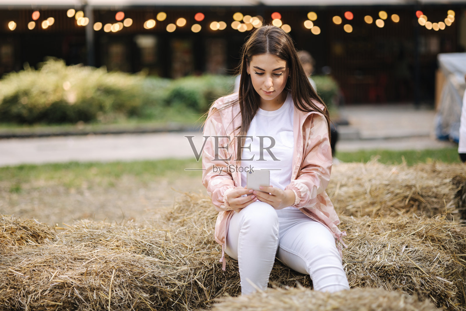 漂亮的女人坐在干草长凳上用电话。白衣美女放松。圣诞彩灯背景照片摄影图片