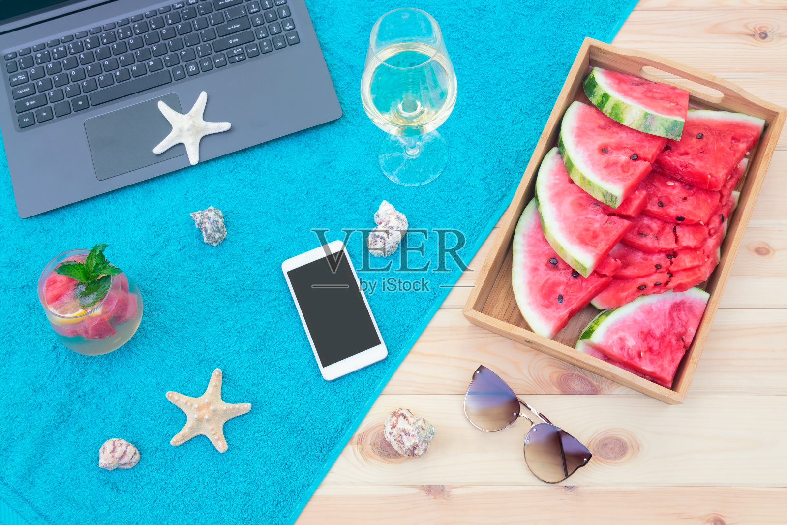 笔记本电脑,智能手机,海星,贝壳,白葡萄酒,西瓜,西瓜柠檬水和太阳镜在木制表面毛巾。前视图。照片摄影图片