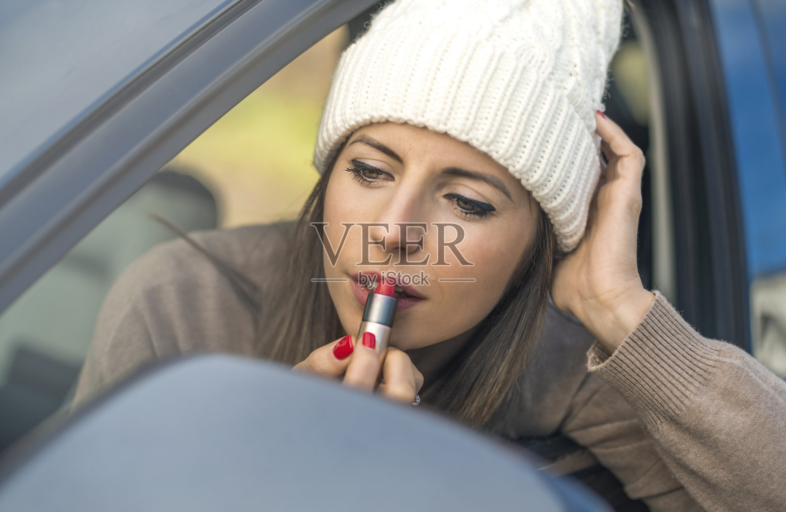 一个戴着白色羊毛帽的小女孩,一边看着汽车的后视镜,一边把嘴唇涂成红色。照片摄影图片