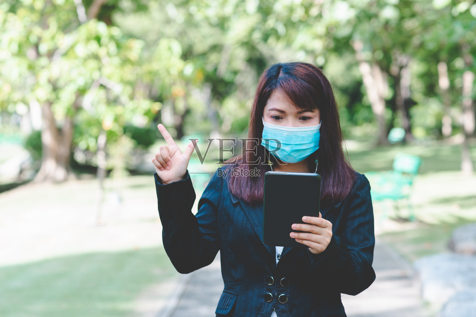 成功的商业女性戴着口罩使用智能手机平板电脑在屏幕上打字与应用设备在公园与自然背景。由于冠状病毒或covid-19,商业进入新常态。照片摄影图片