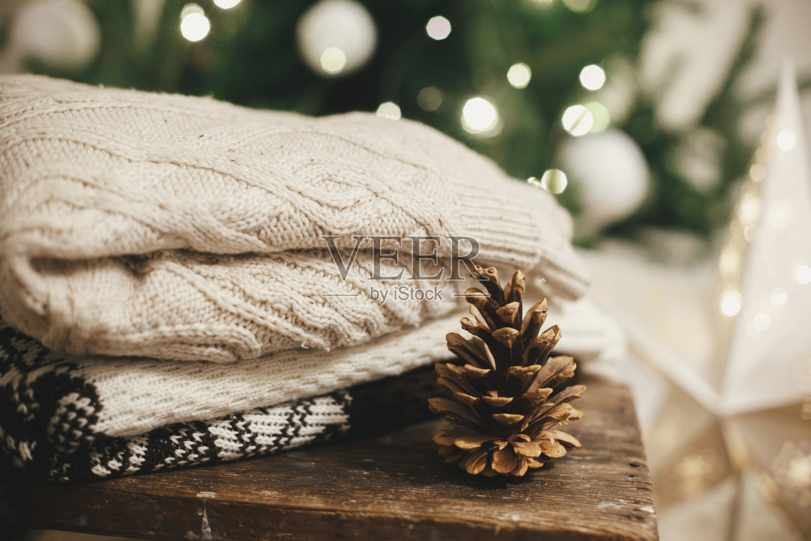 舒适的针织毛衣在乡村木材与松果背景圣诞树在灯光。一堆时尚的毛衣在节日装饰的斯堪的纳维亚房间。大气冬天照片摄影图片