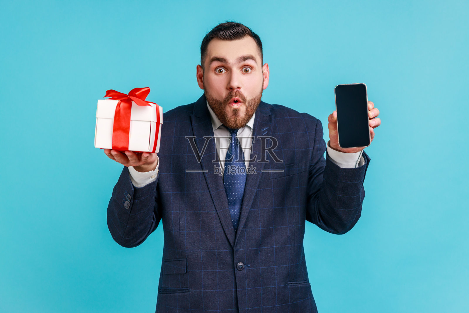 哇,移动用户的礼物奖励!年轻的成年男子穿着深色西装,手持礼品盒和带有空白显示的手机,用于广告照片摄影图片