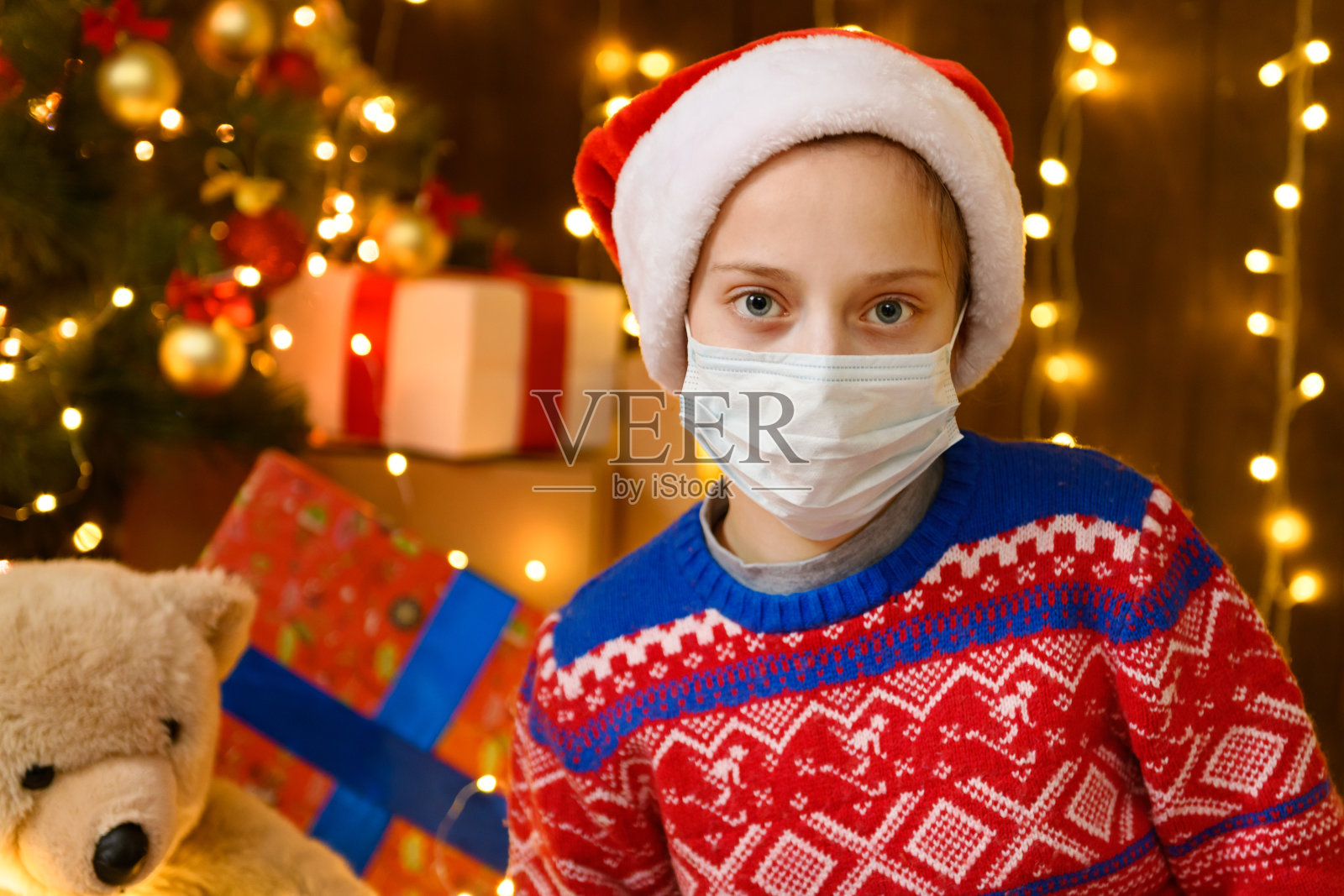 小女孩作为圣诞老人的助手,在新年的装饰。戴上防护口罩,抵御病毒,冠状病毒。节日彩灯和许多礼物,一棵优雅的圣诞树和玩具。照片摄影图片