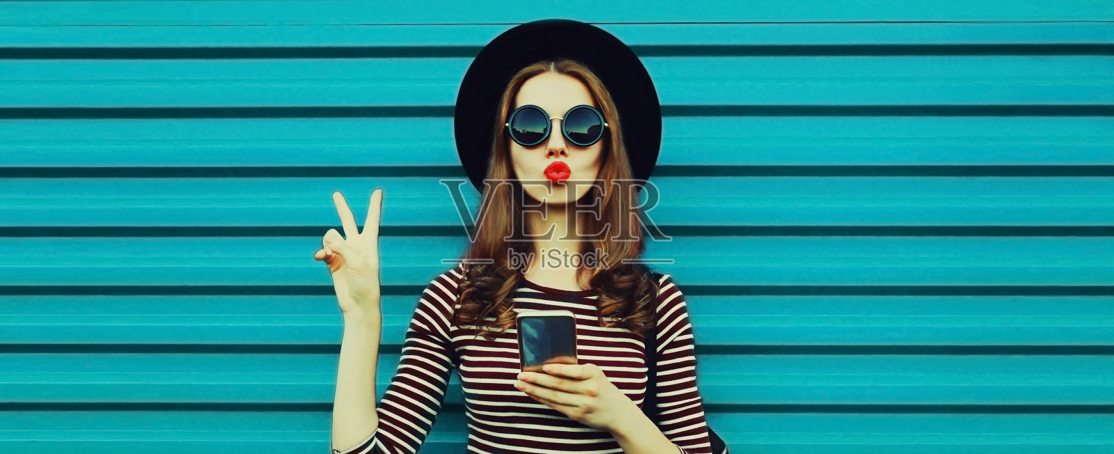 时尚年轻女模特的肖像,戴着黑色圆帽,拿着智能手机,背景是蓝色照片摄影图片