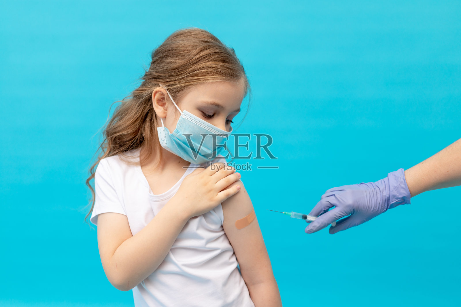 一个小女孩在接种疫苗后,手臂上带着贴片,戴着蓝色背景的医用防护口罩,护士的手拿着注射器,空白的文字,疫苗的概念照片摄影图片