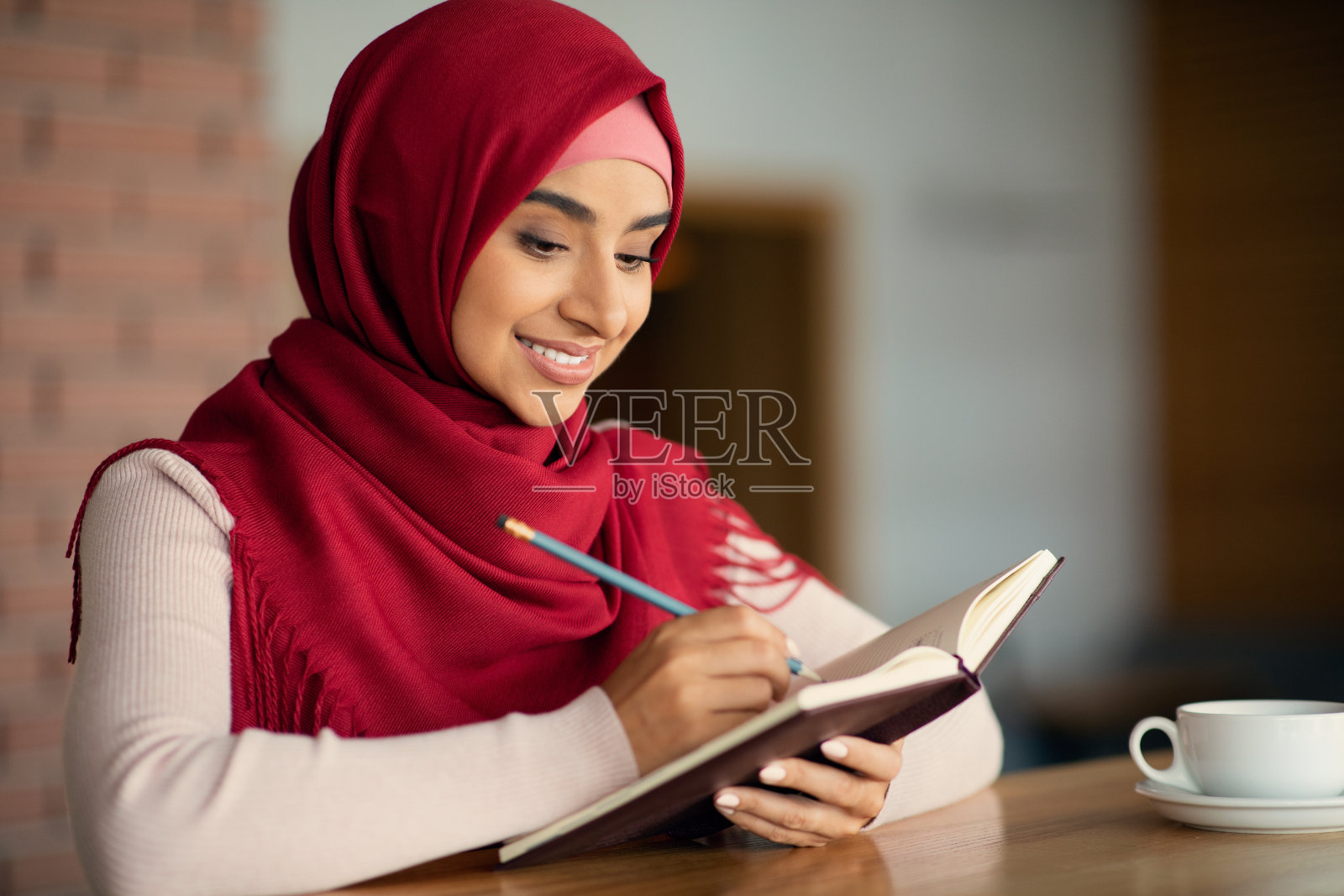 微笑的年轻戴头巾的穆斯林女性在咖啡馆写笔记照片摄影图片