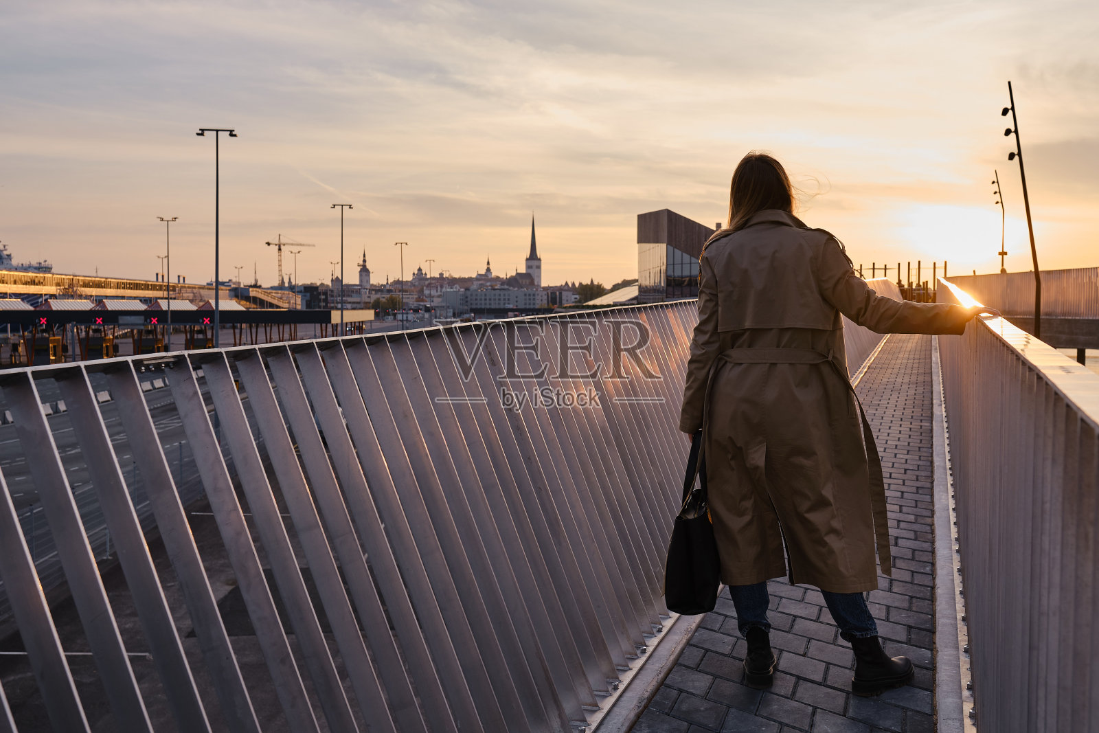 一个穿着风衣的女孩站在城市里一座现代化的桥上看日落。游客在欧洲漫步。城市的生活方式照片摄影图片