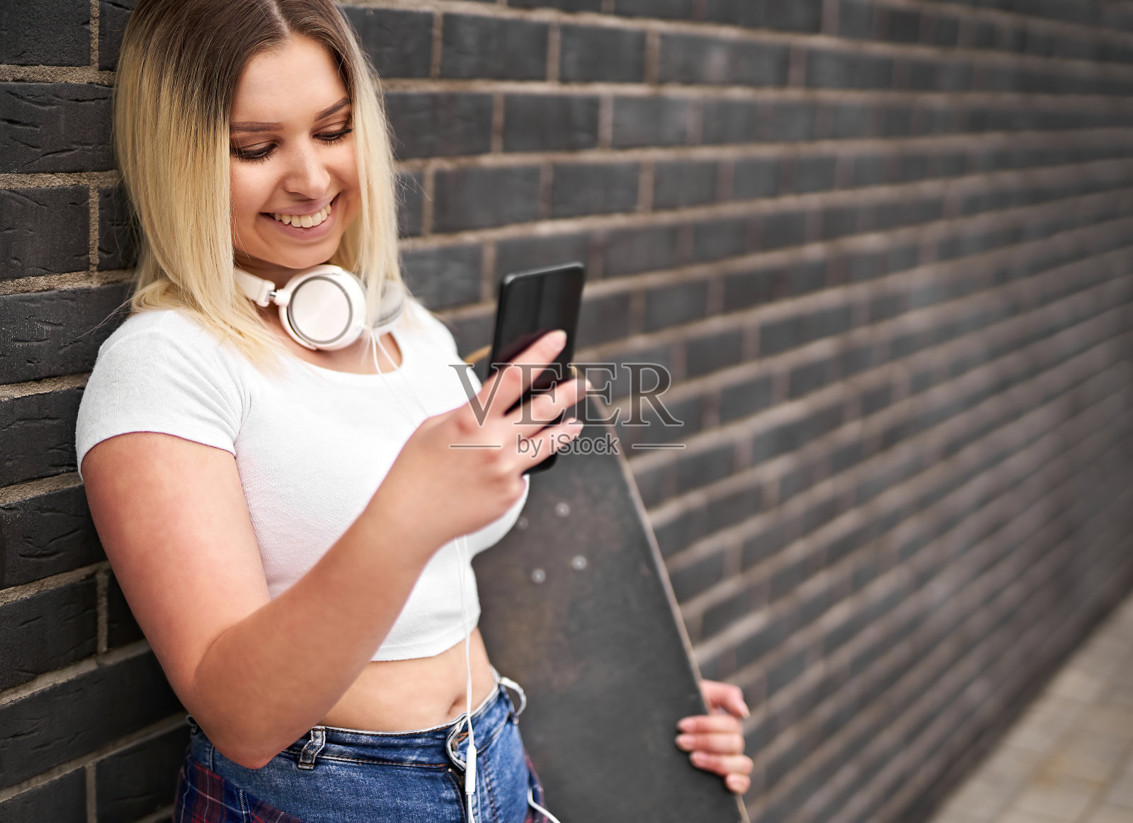 快乐年轻开朗的千禧女学生站在一堵黑砖墙前,拿着滑板用智能手机聊天-人们的生活方式和技术概念与青少年照片摄影图片