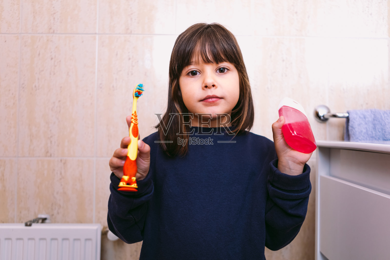 小女孩穿着深蓝色的运动衫,拿着牙刷和牙膏在浴室里。刷牙、卫生与童年观念。照片摄影图片