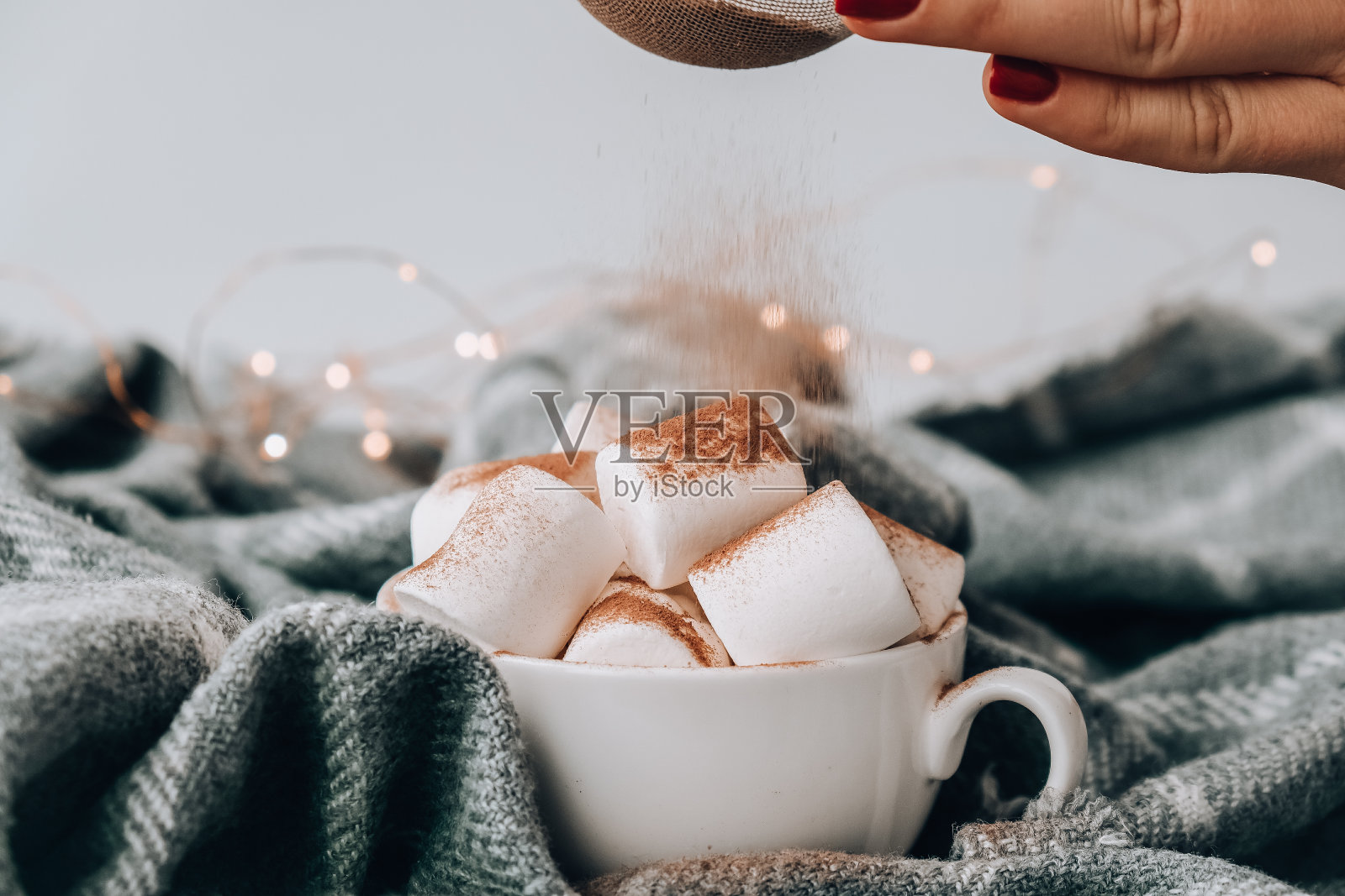 在热巧克力上加入可可粉,白色杯子里放棉花糖,毛衣为背景。舒适的圣诞灯照片摄影图片