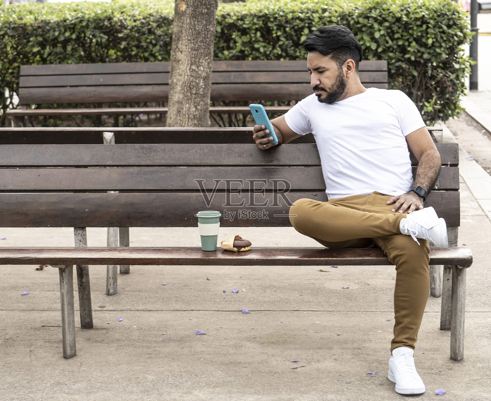 拉丁人坐在广场长凳上看着手机,喝着咖啡,吃着甜甜圈照片摄影图片