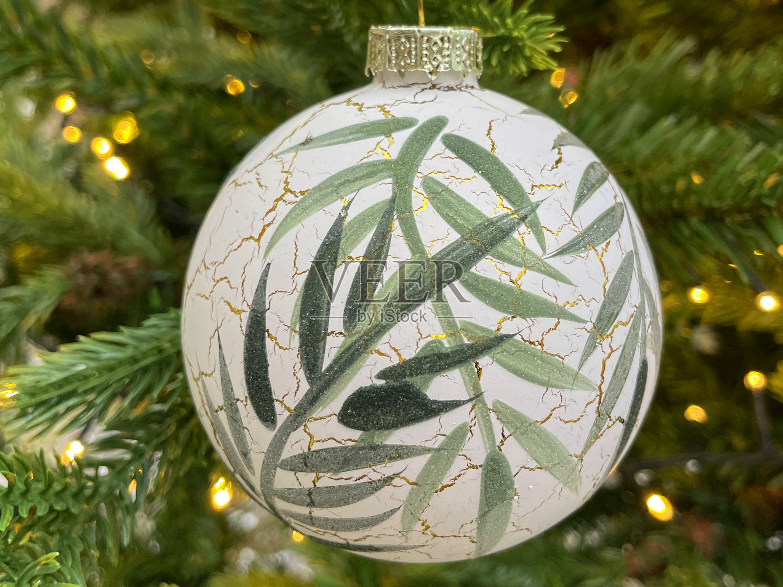 个人特写图像,白色的小装饰物用绘成绿色的竹叶装饰,挂在人造的圣诞树树枝上,绿色的云杉针,模糊的白色精灵灯背景照片摄影图片