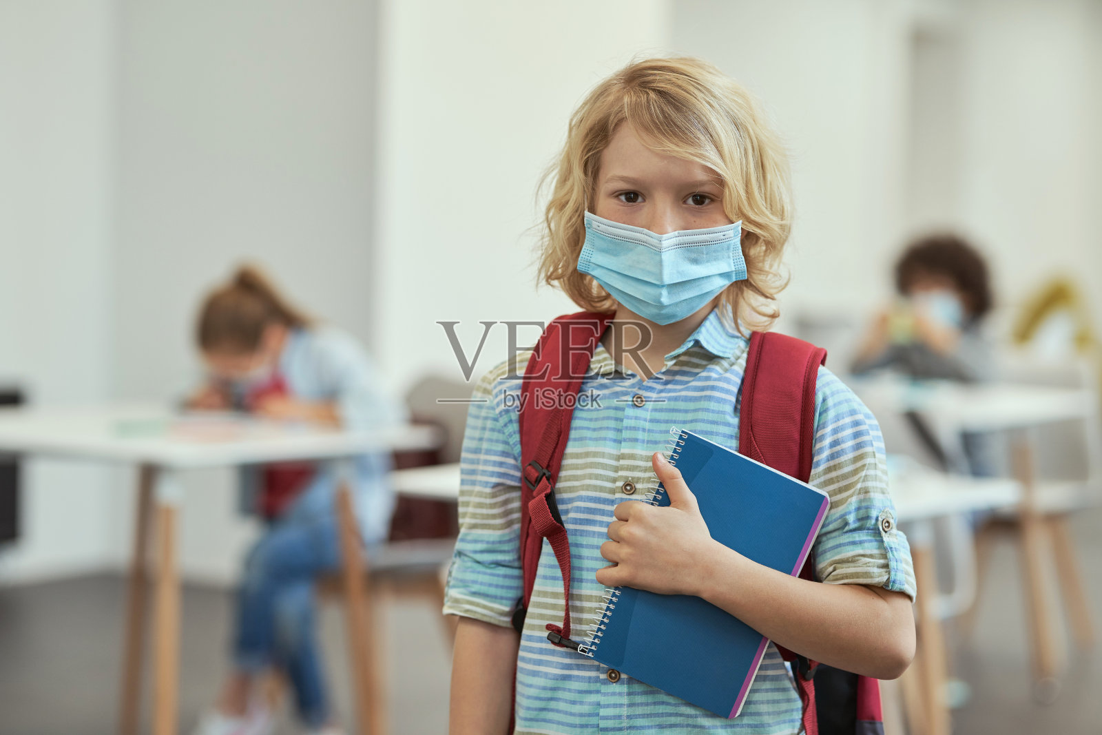 为了防止covid - 19的传播,戴着口罩的安静小男孩在教室里摆姿势,看着相机,拿着笔记本照片摄影图片