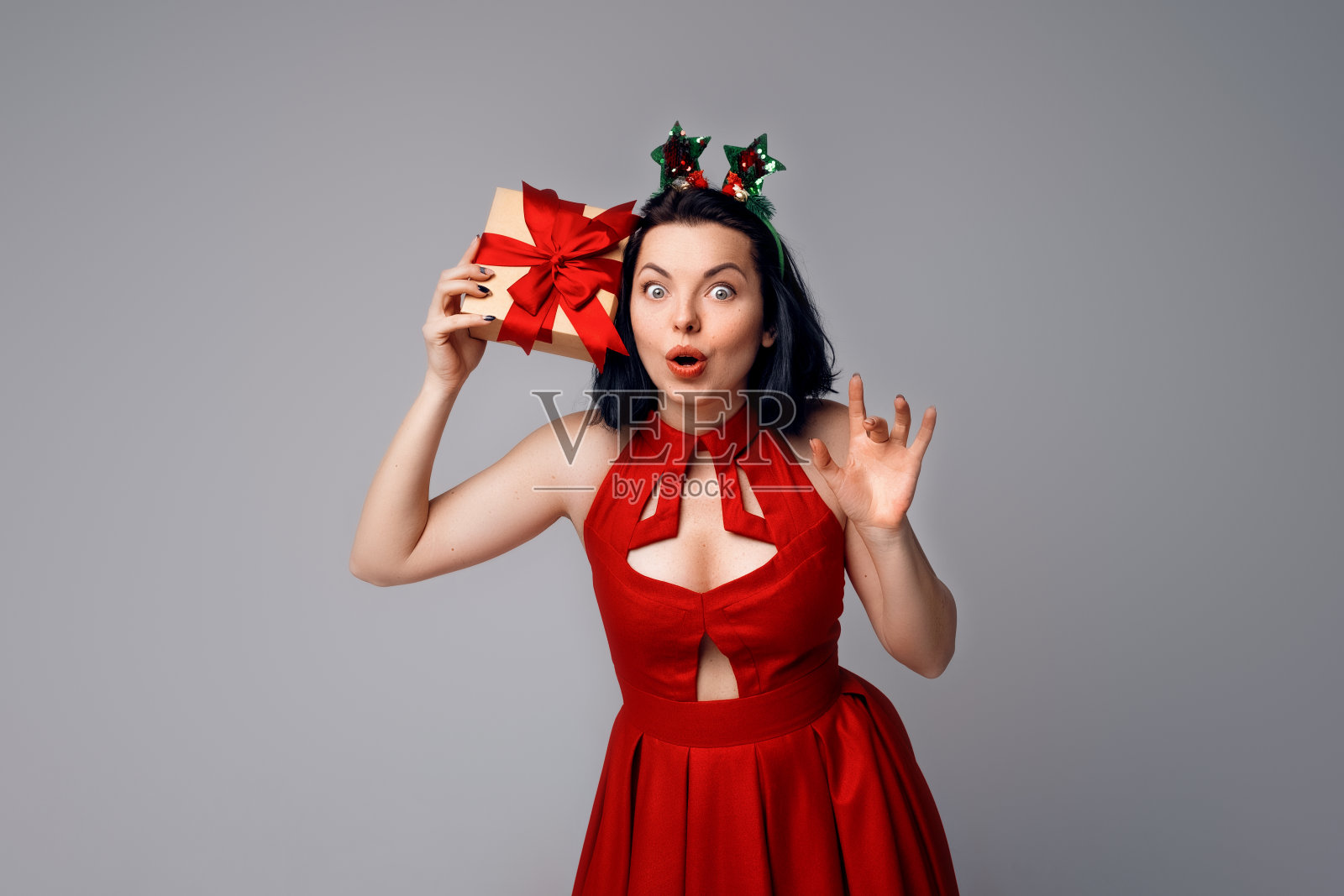 好奇的年轻女子想知道礼品盒里面是什么。在灰色背景上穿红裙子的快乐女孩。假日,庆典和生活理念照片摄影图片