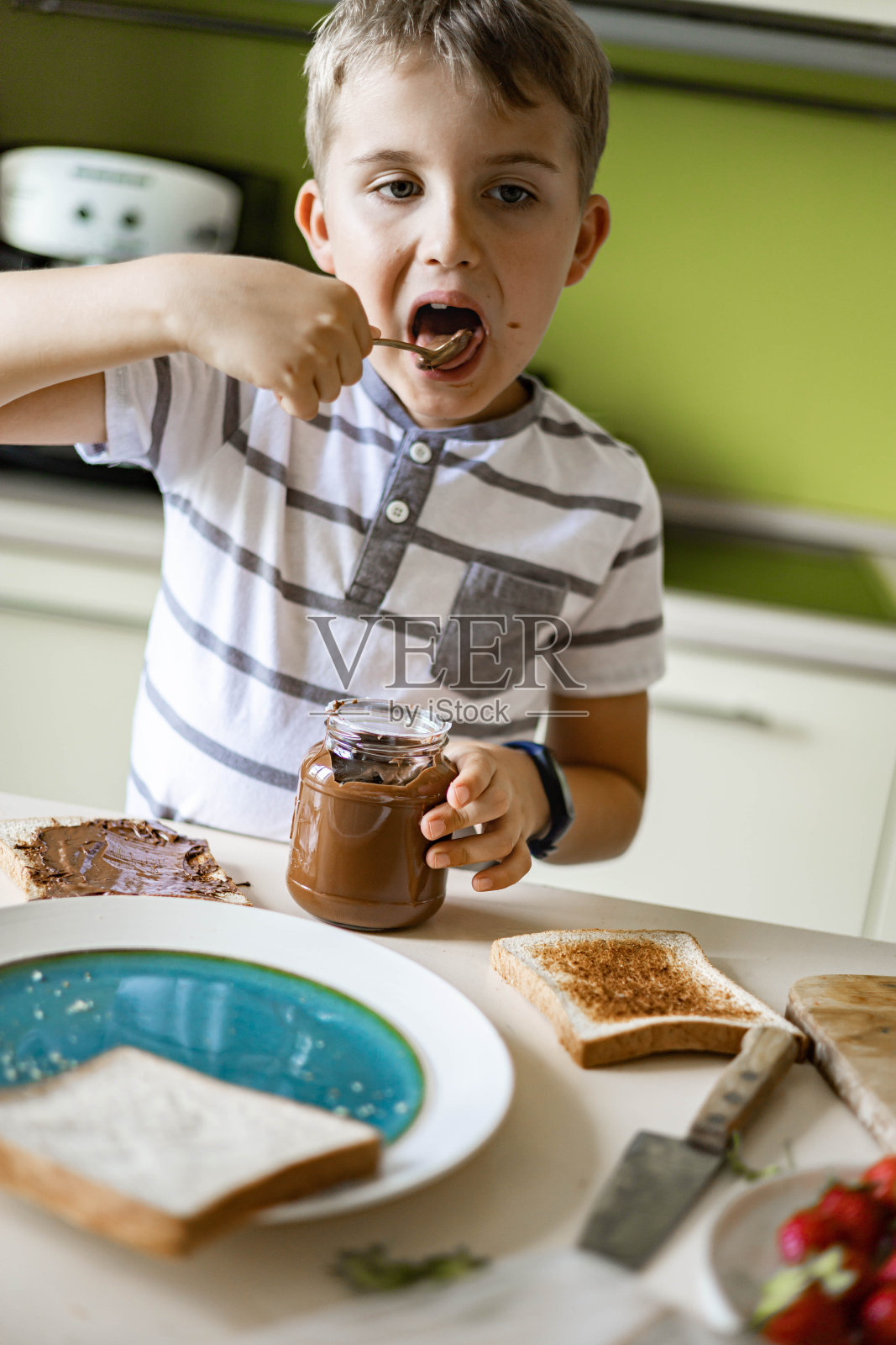 一个小男孩吃着从罐子里取出的巧克力意大利面,用勺子在现代厨房的桌子上做三明治照片摄影图片
