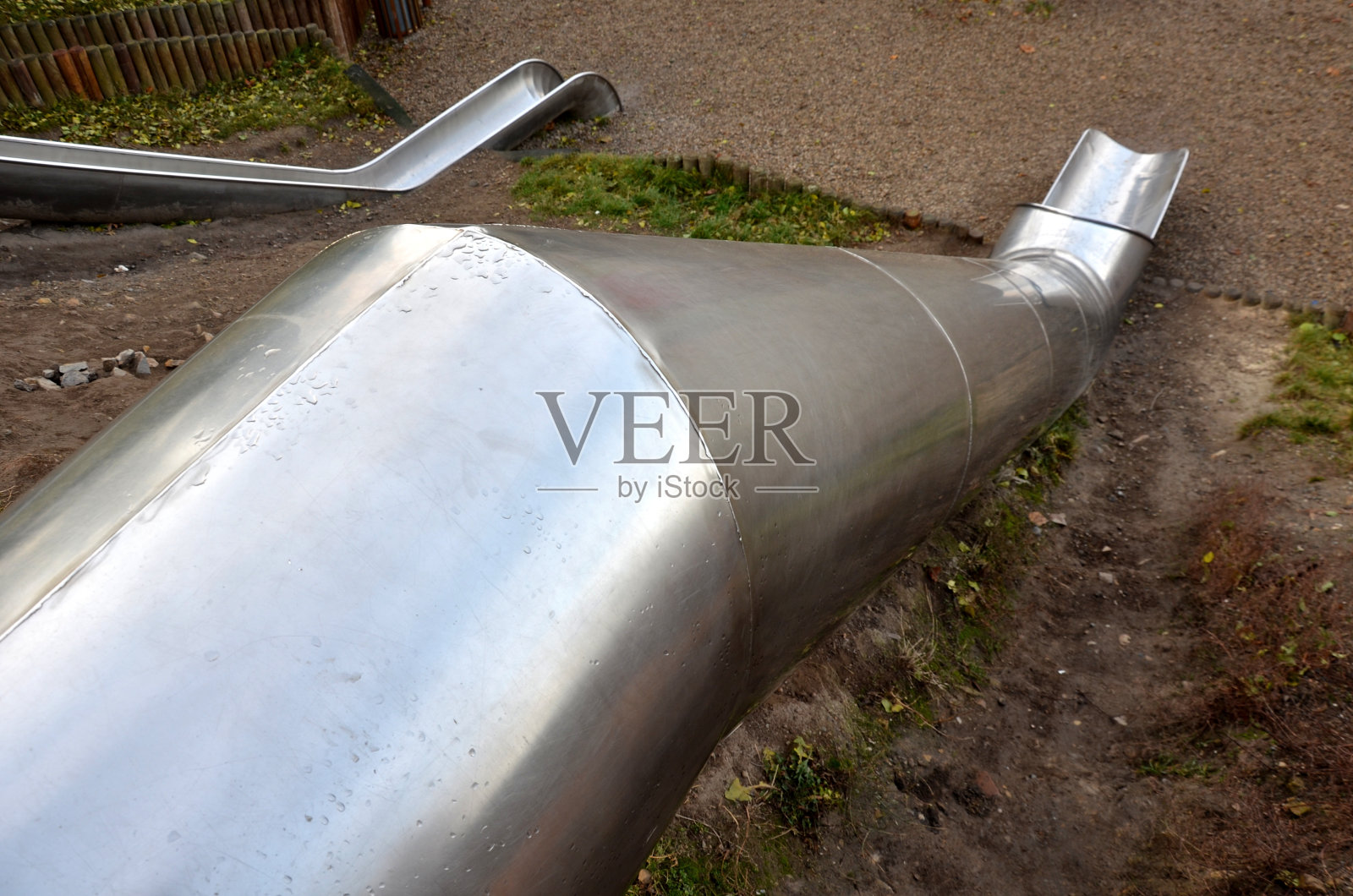 球场上有不锈钢滑梯,周围有草山和木制栅栏。大型光滑管滑动儿童与冲击格子橡胶垫。管子的末端是一个开口槽照片摄影图片