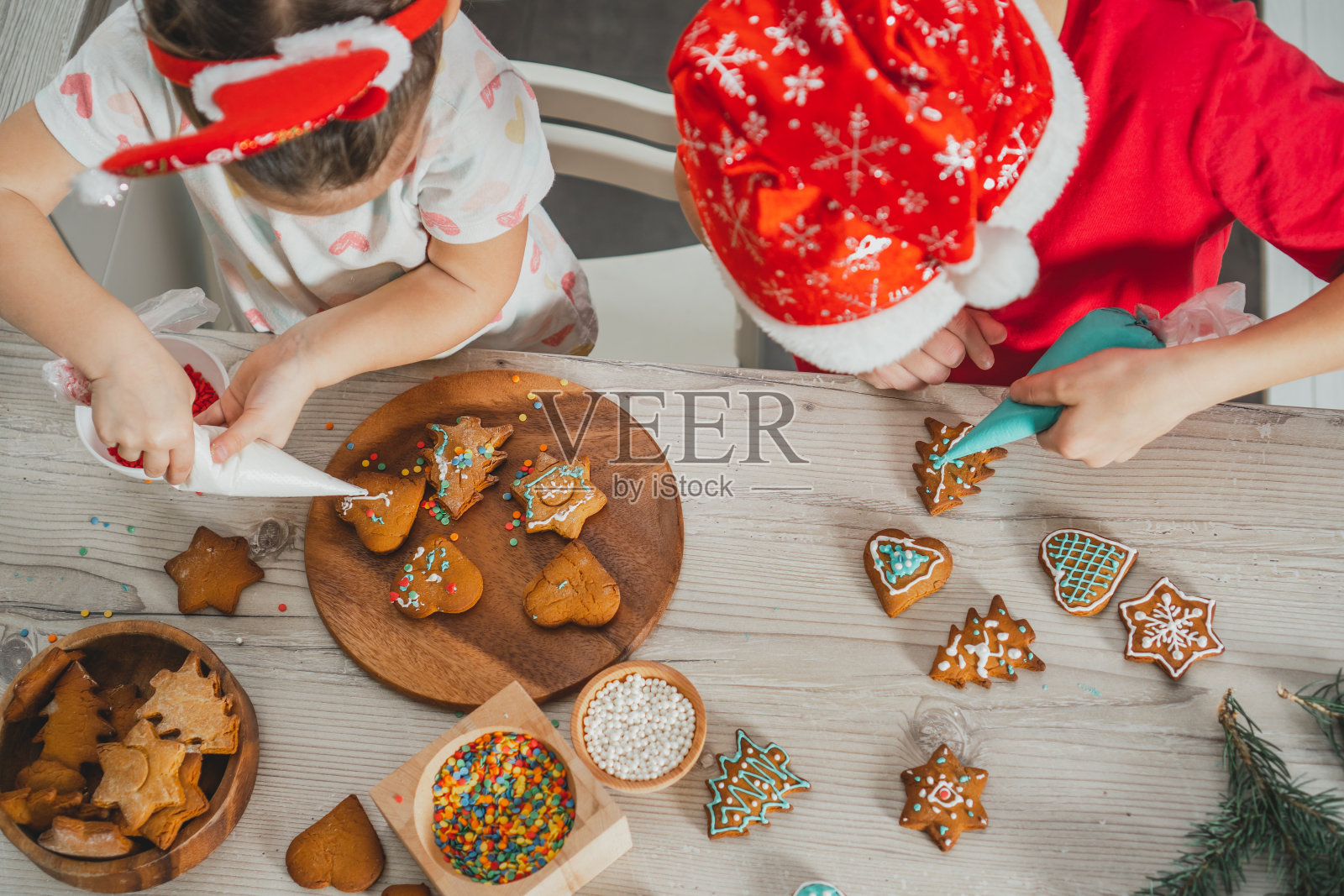 3岁的女孩和8岁的男孩戴着圣诞帽用糖霜装饰姜饼饼干。圣诞厨房里的兄弟姐妹。前视图。照片摄影图片